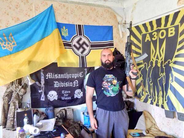 A man poses in front of flags representing Ukrainian Neo-Nazi groups, including the aptly-named Misanthropic Division. 