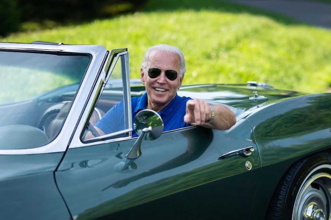 Biden smiling and pointing at you from the driver's seat of a convertible sports car. 