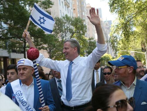 De Blasio has both hands in the air, one waving a flag of Israel as he marches in a parade