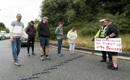 a police officer points to spike strips while talking to a man holding a sign reading "The government are taxing us into poverty"