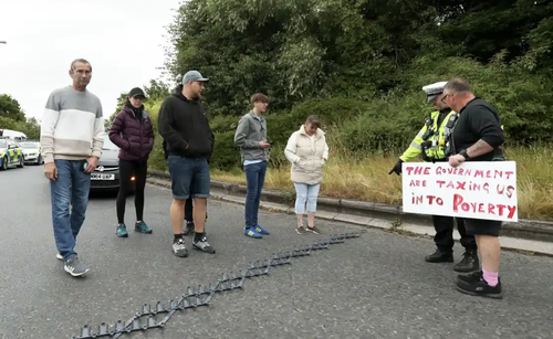 a police officer points to spike strips while talking to a man holding a sign reading The government are taxing us into poverty