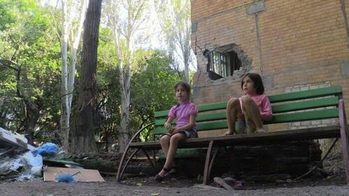 Donbas children sitting in front of a damaged building